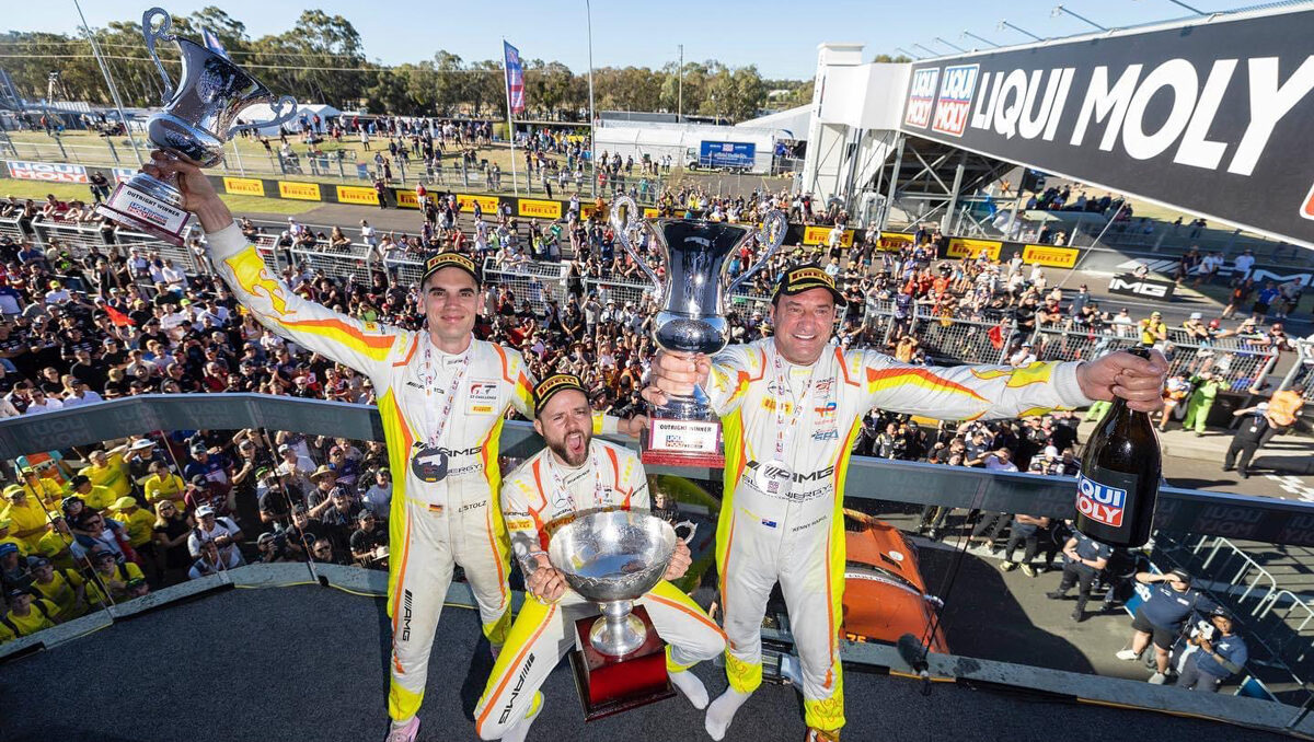 Three race car drivings with a large trophies celebrating in front of a large crowd