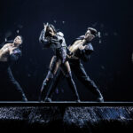 Eurovision dancers wearing black outfits on a dark stage illuminated by spotlights