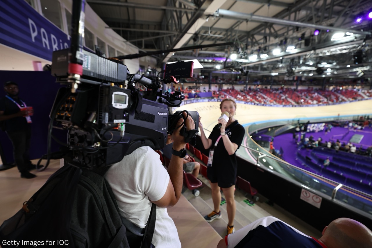 A woman reports in front of a sporting arena while a cameraman films