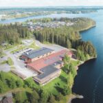 Aerial view of a large lakeside building complex with red and black roofs, solar panels, and surrounding greenery