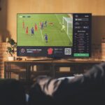 Two people sit on a sofa watching a football game on a TV