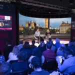 A man and a woman sitting on a stage delivering a presentation to a crowd lit in blue light. Behind the pair, there is an LED screen showing London