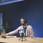 A man in a pink shirt delivers a podcast. He wears headphones and a microphone, phone, water bottle and glass are placed next to him. He uses his laptop to the side