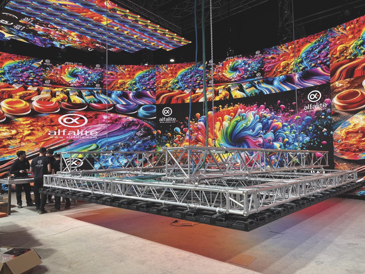 An LED screen with bright colours on it. A metal scaffolding is hanging in front of it and people work off to the side