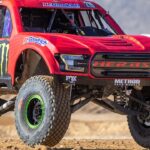 A red off-roading truck with black wheels which is riding over desert terrain