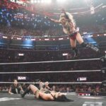 A WWE wrestler jumps from the side of the ring to another wrestler lying on the ground in a packed stadium