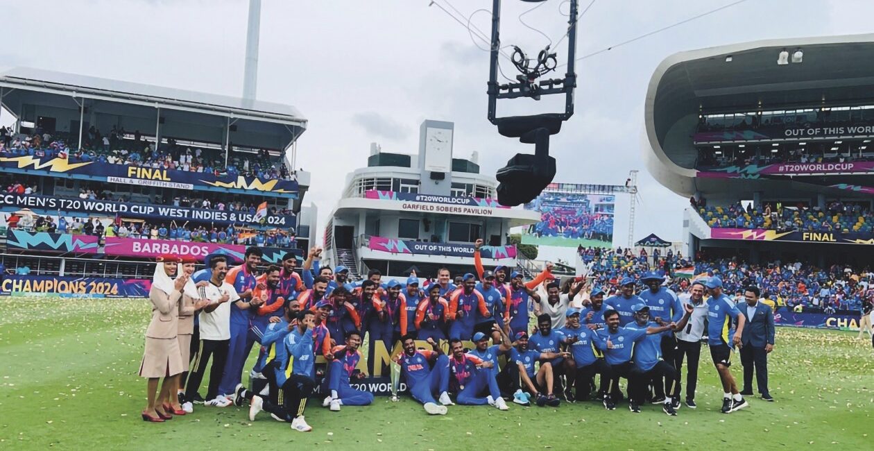 A team sitting on a pitch, smiling at a camera that hangs in the air on a hoist