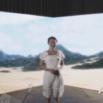 A woman in a white dress standing on a film set in front of a large LED screen showing a desert terrain