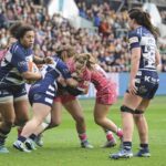 A group of women in red or blue playing rugby in a packed stadium. Three women tackle each other while another runs away with the ball