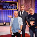 A group of men standing in front of a sign in German on a quiz show