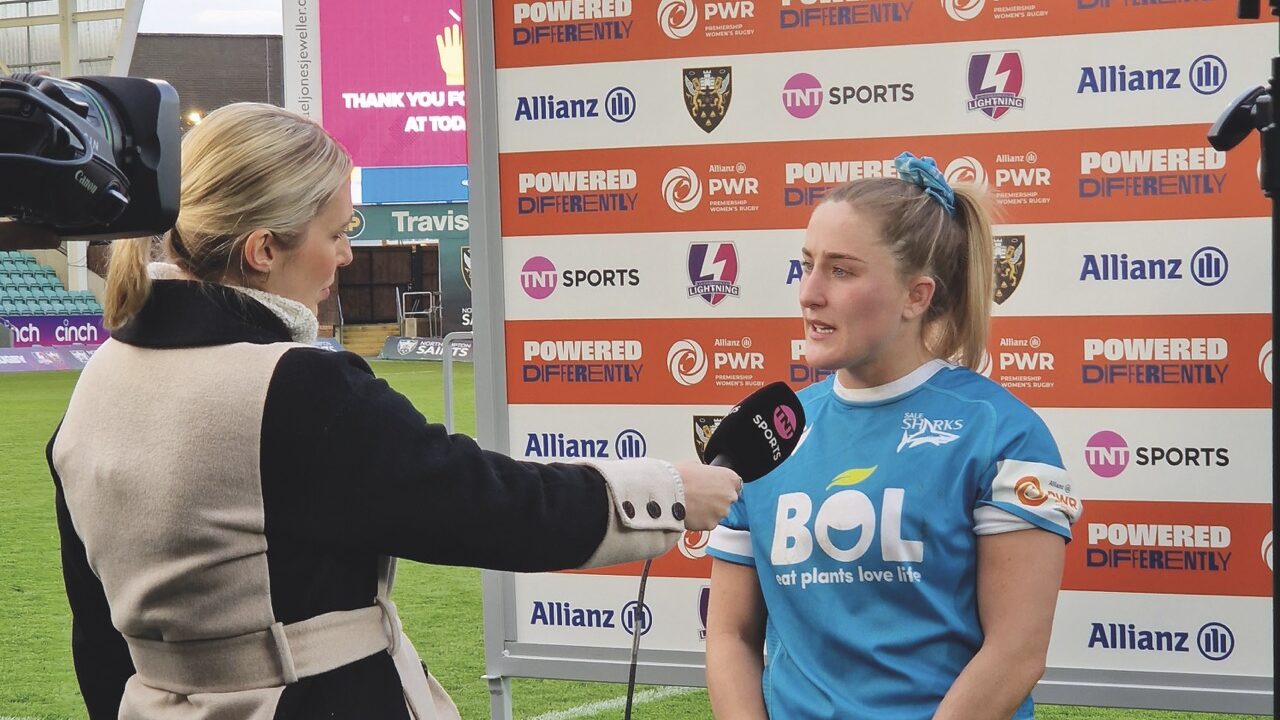 A woman in blue sports clothing being interviewed by a woman wearing a coat on a rugby pitch in front of a display