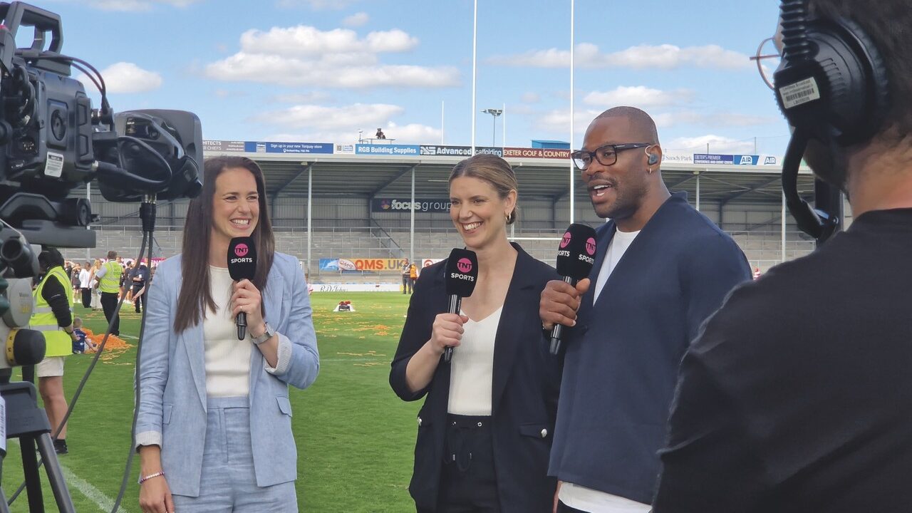 Three people holding large microphones and smiling at a camera. They stand on a rugby pitch