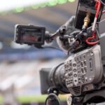 A video camera in the foreground of a blurred football stadium