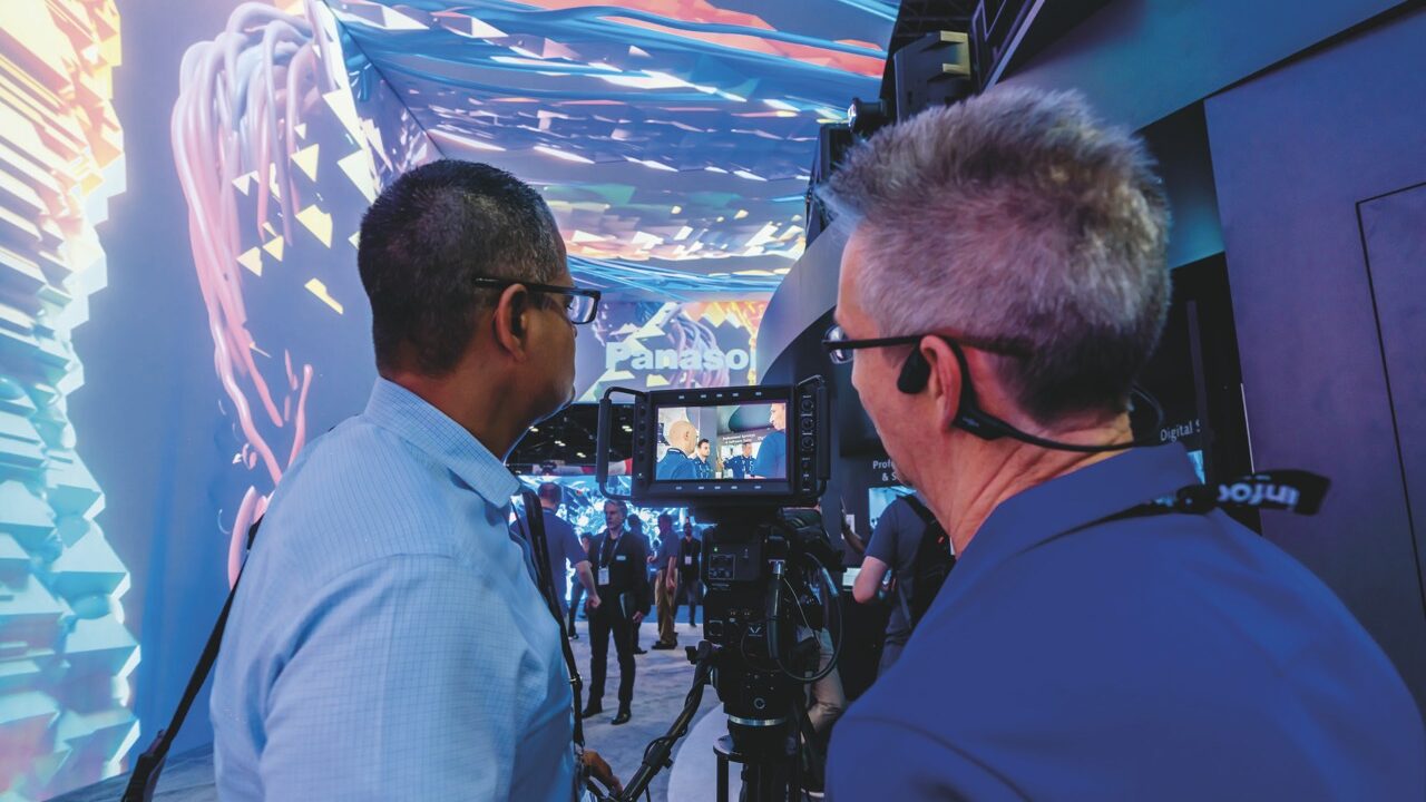 Two men looking at the screen of a camera. They stand in a room with a large projection on the wall and ceiling
