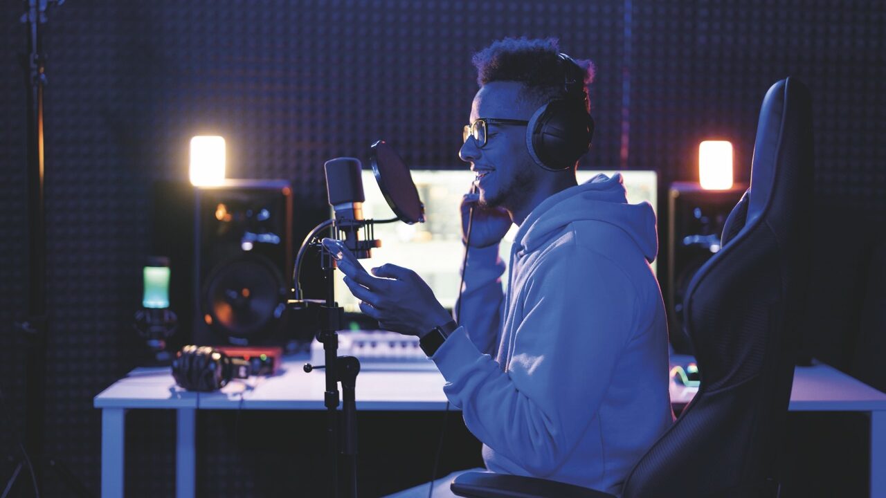 A young man sits at a desk in a dimly lit room. He smiles as he uses a microphone and headphones