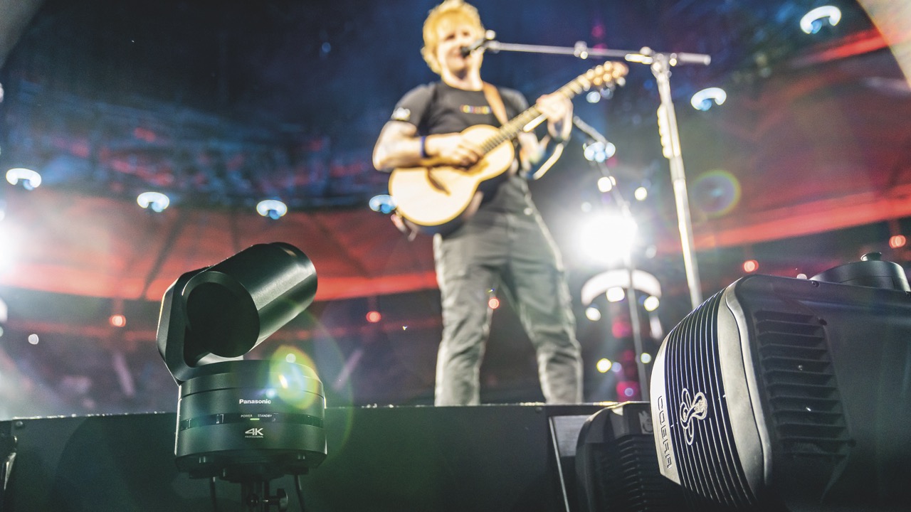 A singer onstage in a blurred background holding a guitar and singing into a microphone. A camera films him from offstage
