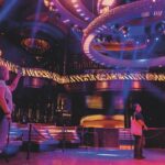 Three people standing in a club setting lit in purple. One takes a photo of the roof. There is seating around the edge of the room that is roped off