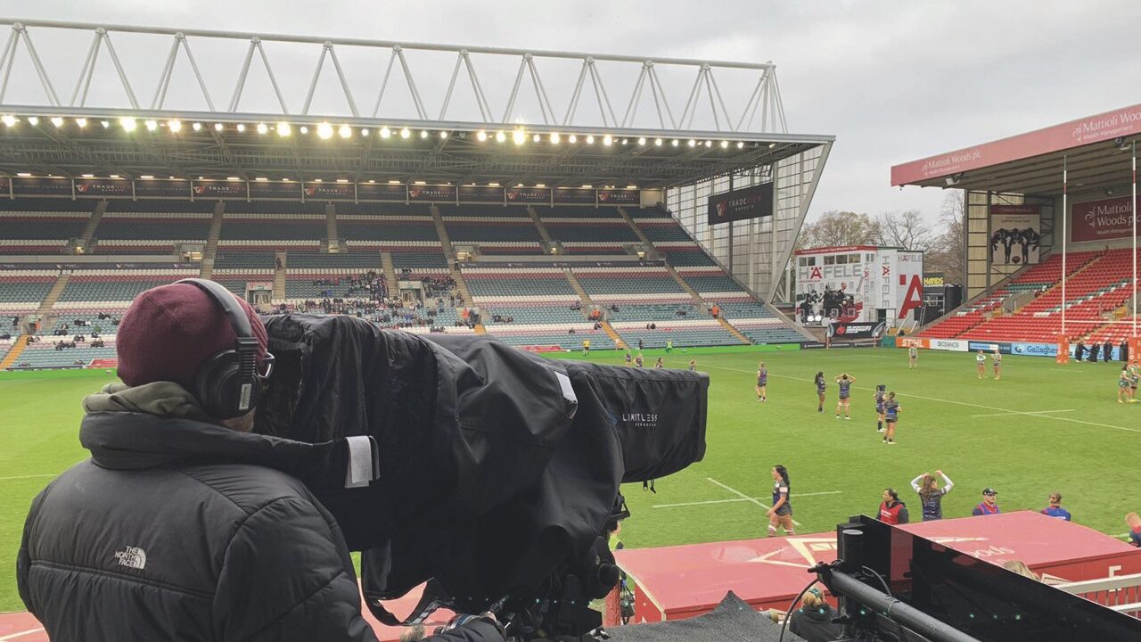 A person with a coat and hat on staring through a camera with a black cover. They film a rugby pitch