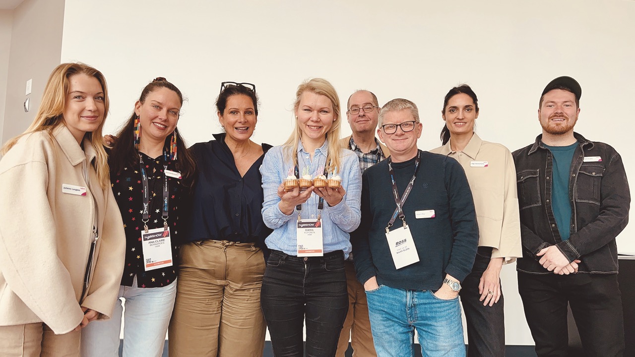 A group of people stand smiling at the camera. Some of them wear large badges around their neck. A woman in the centre holds something flat in her hands