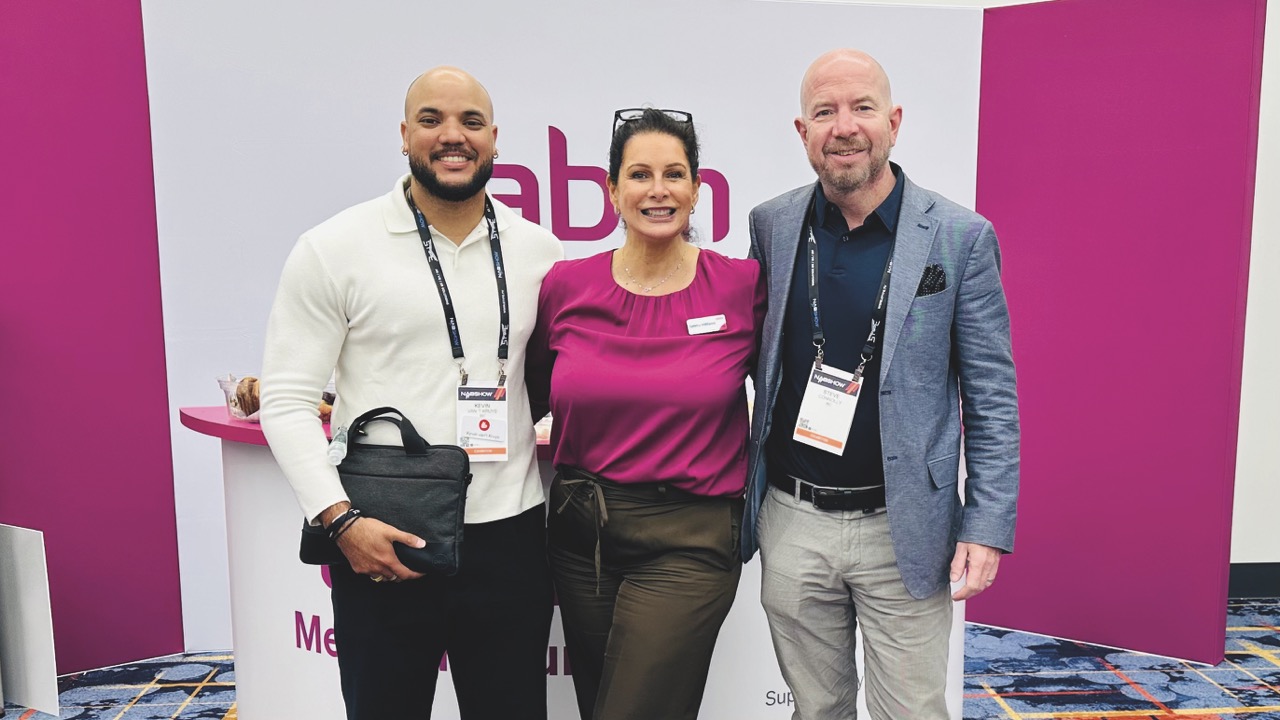 Three people stand in a row with their arms around each other, smiling at the camera. They all wear badges and identification