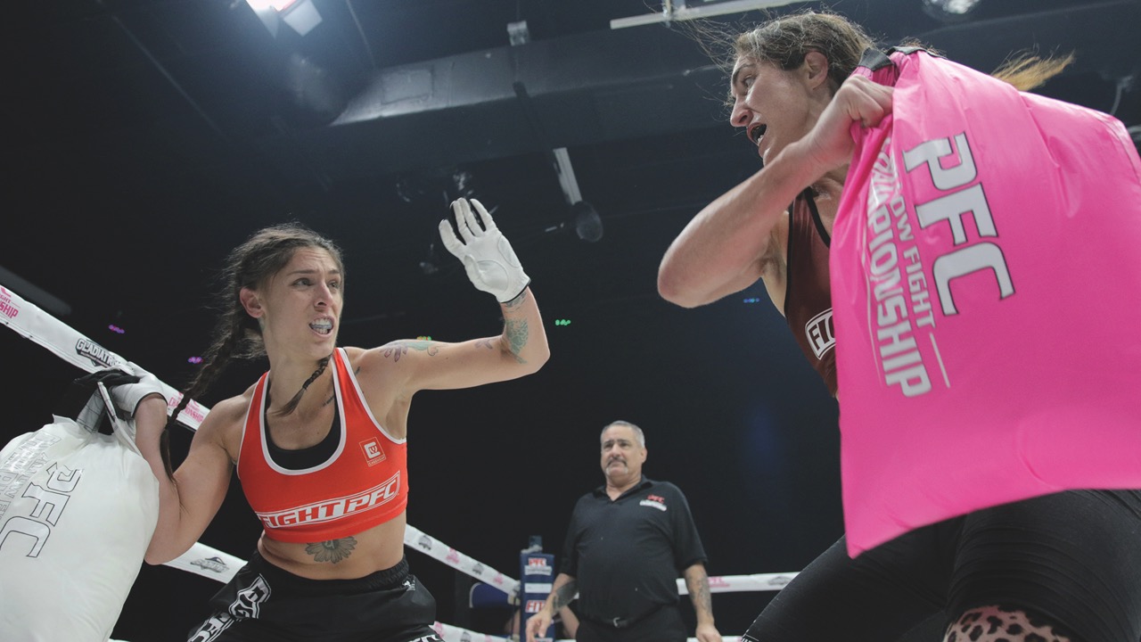 Two women stand in a boxing ring and swing bags at each other. They wear gum shields and one tries to defend with her arm up