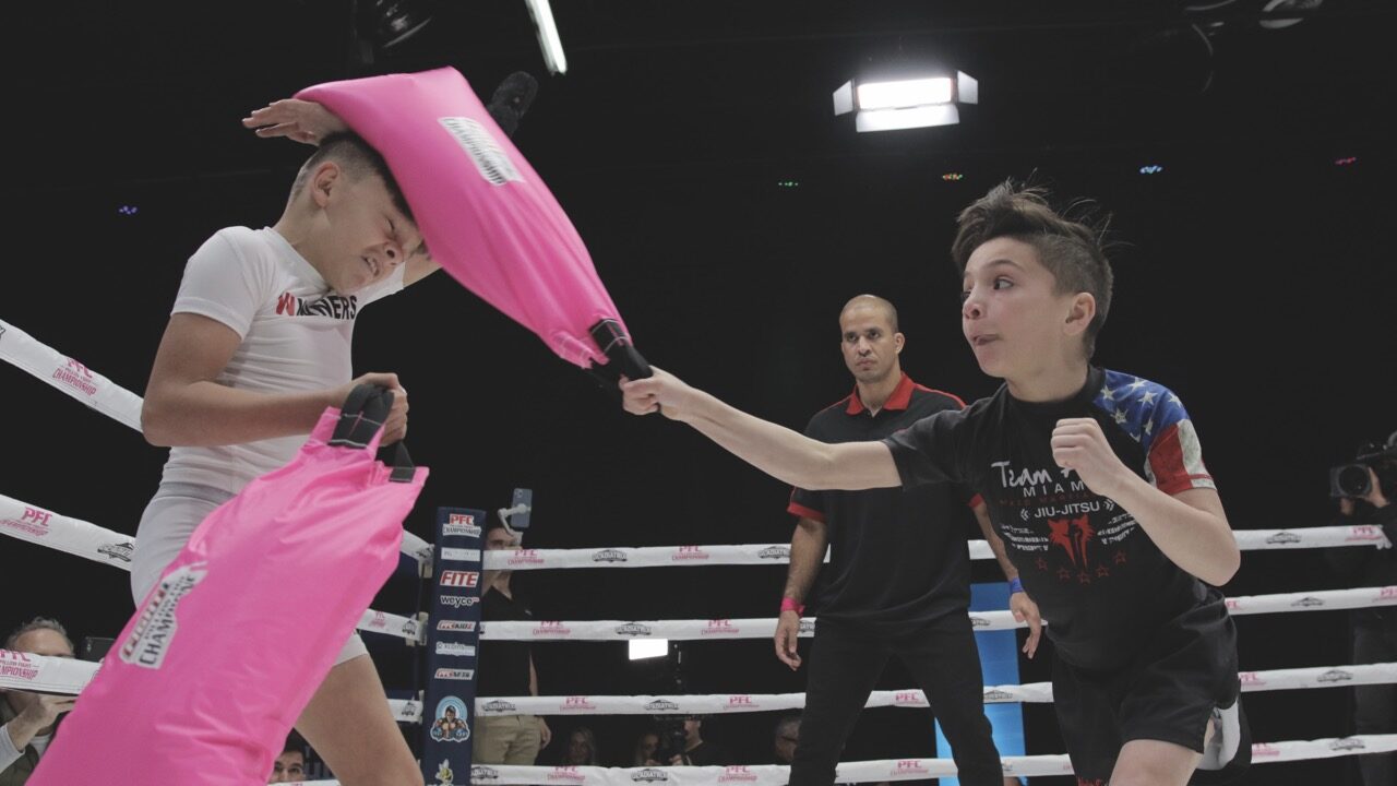 Two children stand in a boxing ring and hit each other with bright pink bags. One boy is being hit in the head