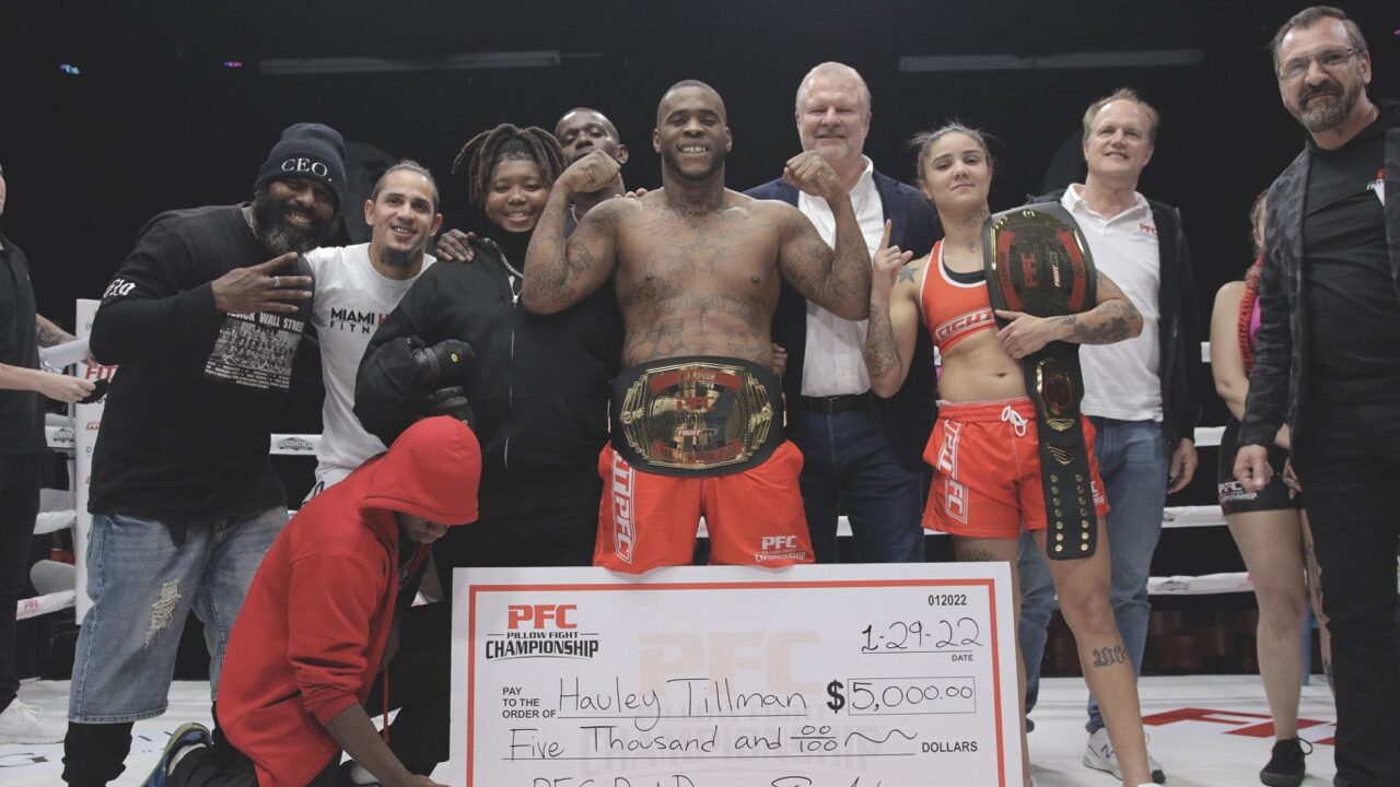 A group of athletes stand in a boxing ring posing in front of a large cheque. Two of the athletes wear large boxing belts