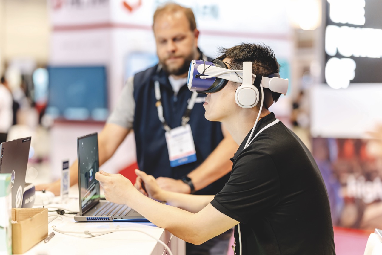 VR Large A young man sitting at a table in front of a laptop. He wears a VR headset and another man stands near him wearing a large badge
