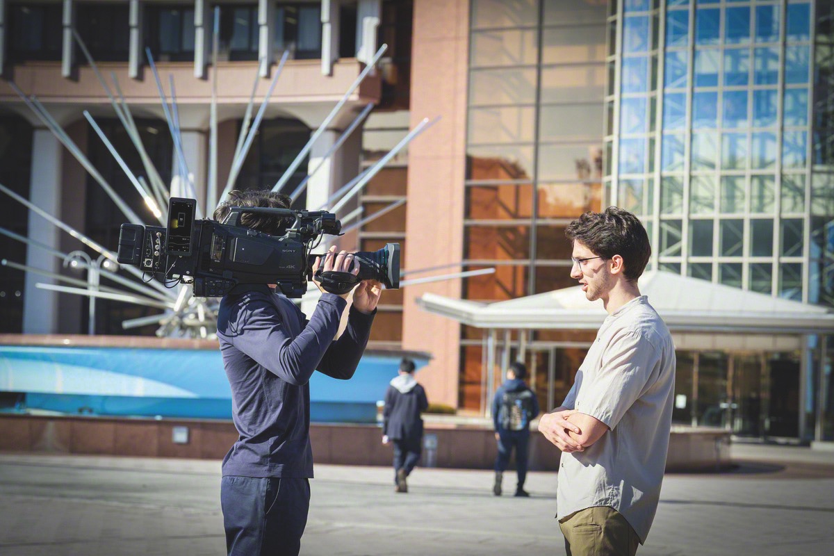 A man standing outside a tall building while another person holds a large camera filming them