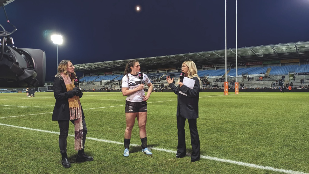Two interviews stand with a woman rugby player on a pitch at nighttime