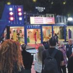 A large warehouse with a mock building inside. A crowd stands around the outskirts watching