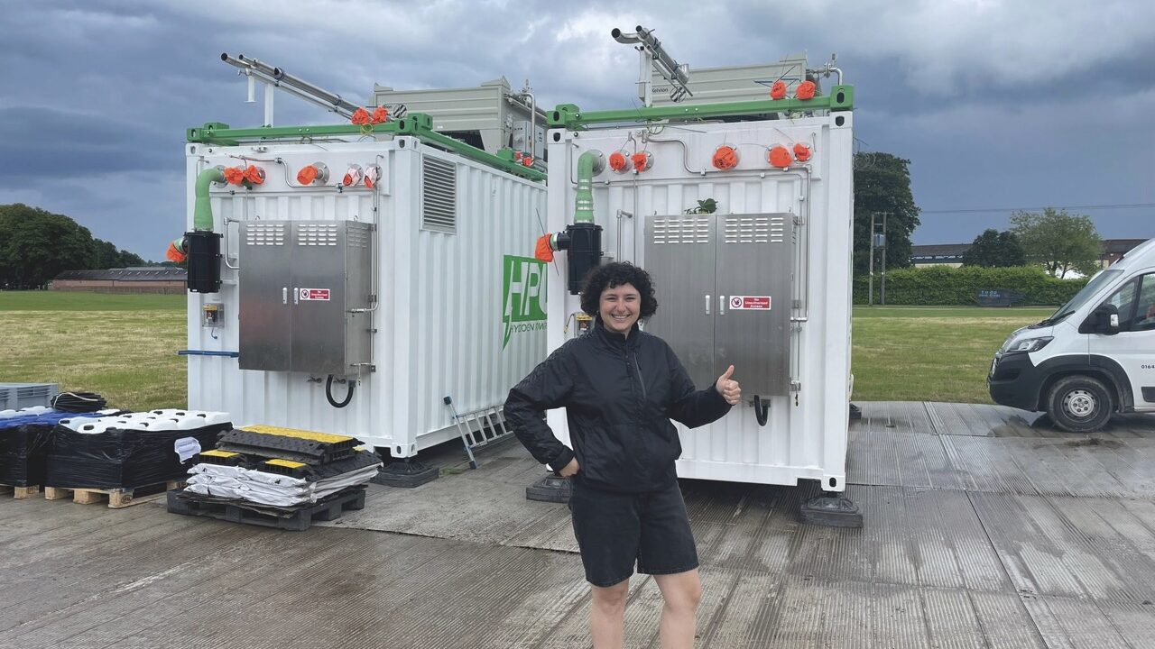 A woman stands outside with her thumb up. She stands next to two large white containers