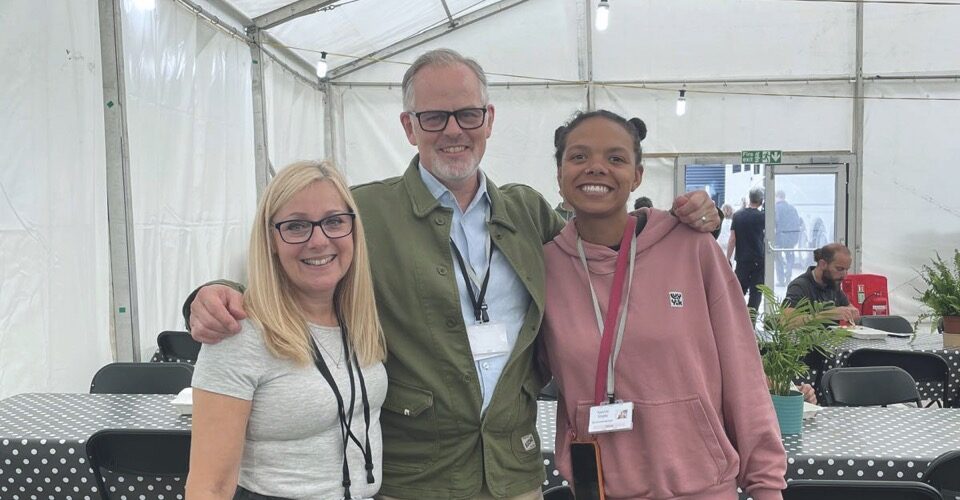 Three people smiling with their arms around each other. They stand inside a white tent