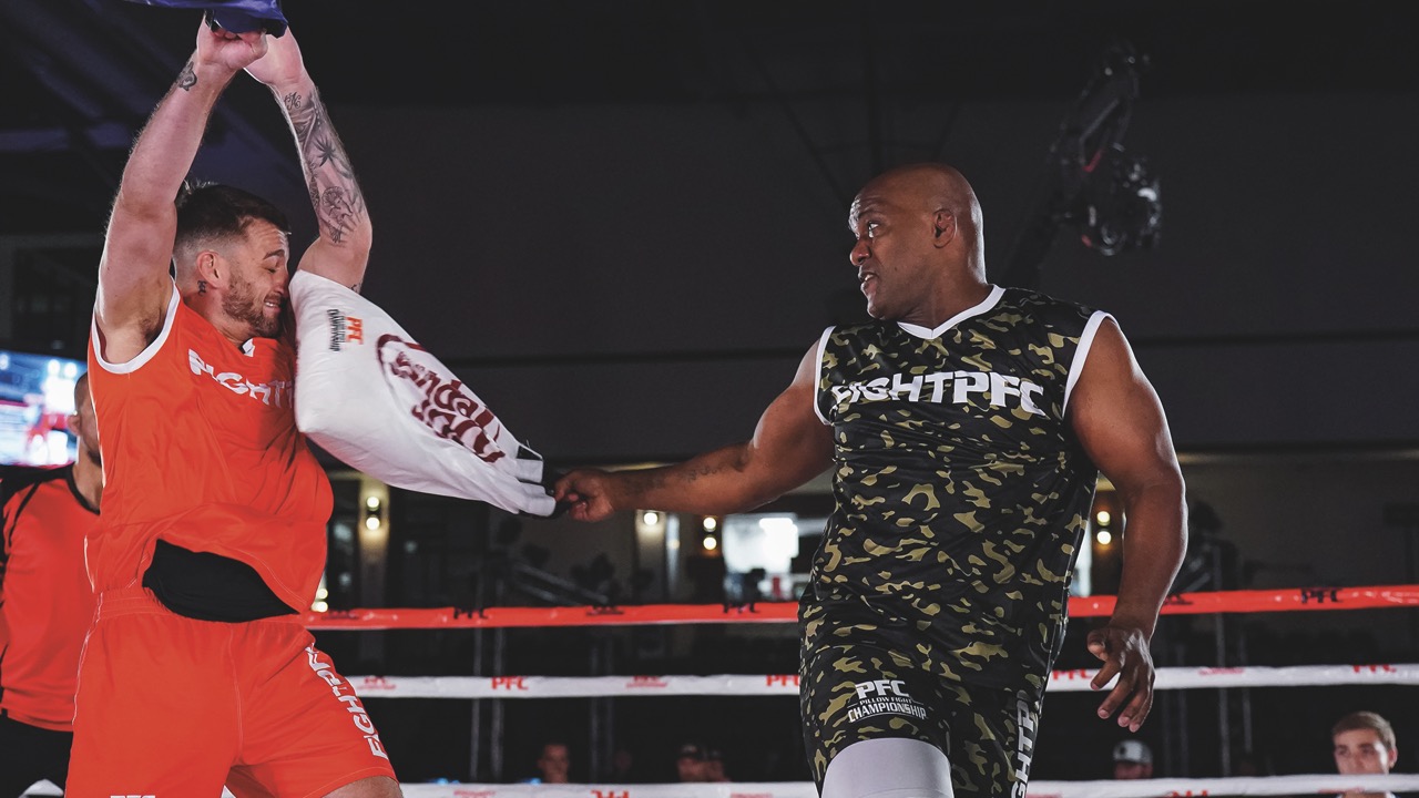 Two men stand in a boxing ring. One wearing red lifts his arms up while being hit by a man holding a white bag