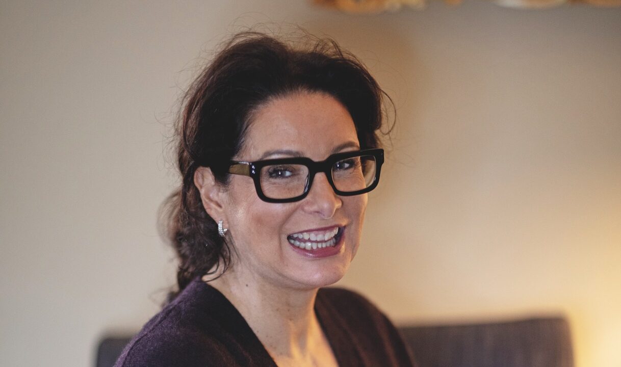 A woman wearing glasses sitting in a room, smiling at the camera