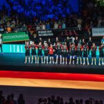A group of people standing in a volleyball arena in a line. A German flag is displayed on the arena’s floor