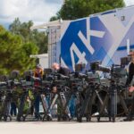 A line of cameras outside, set up in a row next to a large live broadcasting van. A number of people stand and operate the cameras