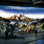A virtual production studio showing a person up a mountain against a mountainous backdrop. People stand in a dark foreground filming the scene
