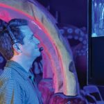 A man wearing a headset looks up at a screen in a dark blue room