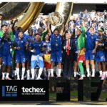 A football team standing on a pitch and celebrating. One teammate holds a silver plate above his head