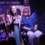 A woman stands onstage at the Women in Media conference, holding a microphone with a panel sat behind