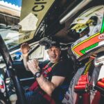 Two people sit in a race car. One wears a helmet, while the other holds audio equipment